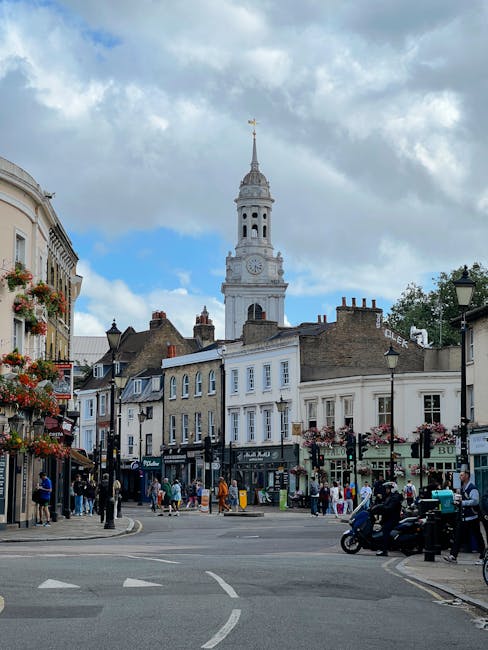A busy street scene in Clapham with a prominent white clock tower with a weather vane on top, situated behind a row of multi-storey buildings that have shopfronts and residential windows, some adorned with flower baskets. The street is filled with pedestrians walking on the sidewalk, some carrying shopping bags, and vehicles including motorcycles and a small car. In the foreground, there is a paved road with white markings and a pedestrian crossing, while black lampposts line the street. The sky above features partly cloudy weather with patches of blue, providing natural daylight that illuminates the scene. This urban setting reflects typical home relocation and furniture transport activities, with the focus on the local community environment and street-level transportation, as would be seen during household removals in the Clapham area. Removal Van Clapham frequently facilitates such house removals and moving services in this vibrant locality.