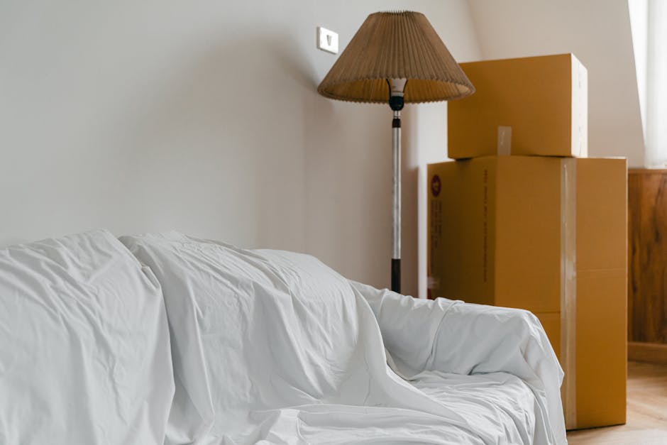 The image shows an interior scene with a white couch partially covered by a protective white sheet, situated against a plain light-colored wall. Behind the couch, there is a tall floor lamp with a brown pleated lampshade standing next to three large cardboard boxes stacked vertically in a corner, some of which have printed markings indicating they are packing materials. The boxes are placed near a wooden piece of furniture, possibly a cabinet or sideboard, and are positioned on the wooden floor. Light from a window on the right side illuminates the scene, creating soft shadows. The environment appears ready for a home relocation process, with packing materials and furniture prepared for transport, as would be typical in professional removals handled by companies like Removal Van Clapham.